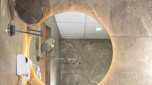 A bathroom with a round mirror, a sink, a hairdryer, and a shower. - Mosaic Connect Ayodhya (Faizabad)