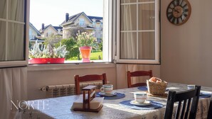 En matplats med ett bord dukat för två personer, ett fönster med gardiner, en klocka på väggen och en utsikt över hus och grönska utanför.