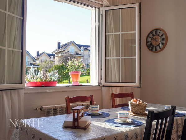 Un coin repas avec une table dressée pour deux personnes, une fenêtre avec des rideaux, une horloge au mur et une vue sur les maisons et la verdure environnante.