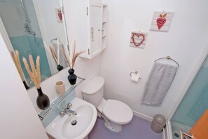 Une salle de bain avec un lavabo, des toilettes et une douche, le tout de couleur blanche. On y trouve également des éléments décoratifs, comme un vase et un porte-serviettes.