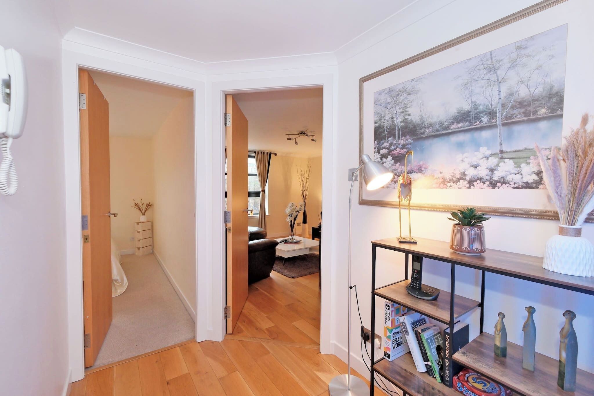 Un pasillo con suelo de madera, pared blanca y una puerta que da a una habitación con sofá negro y una mesita.