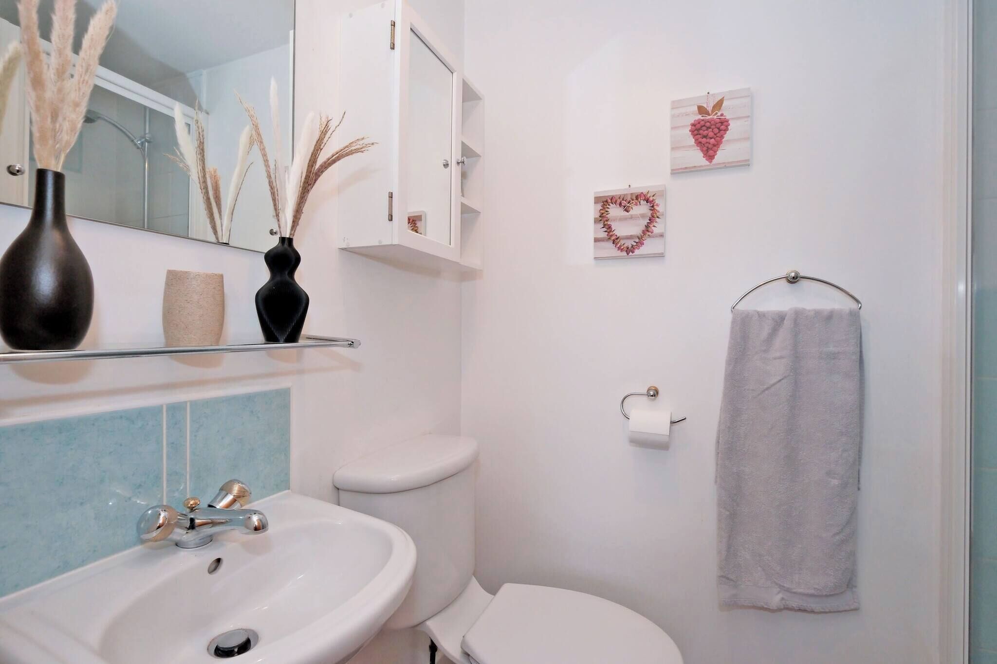 A bathroom with a white sink, toilet, and shower area. There are decorative items on the wall, including a heart-shaped decoration and a vase with dried plants.