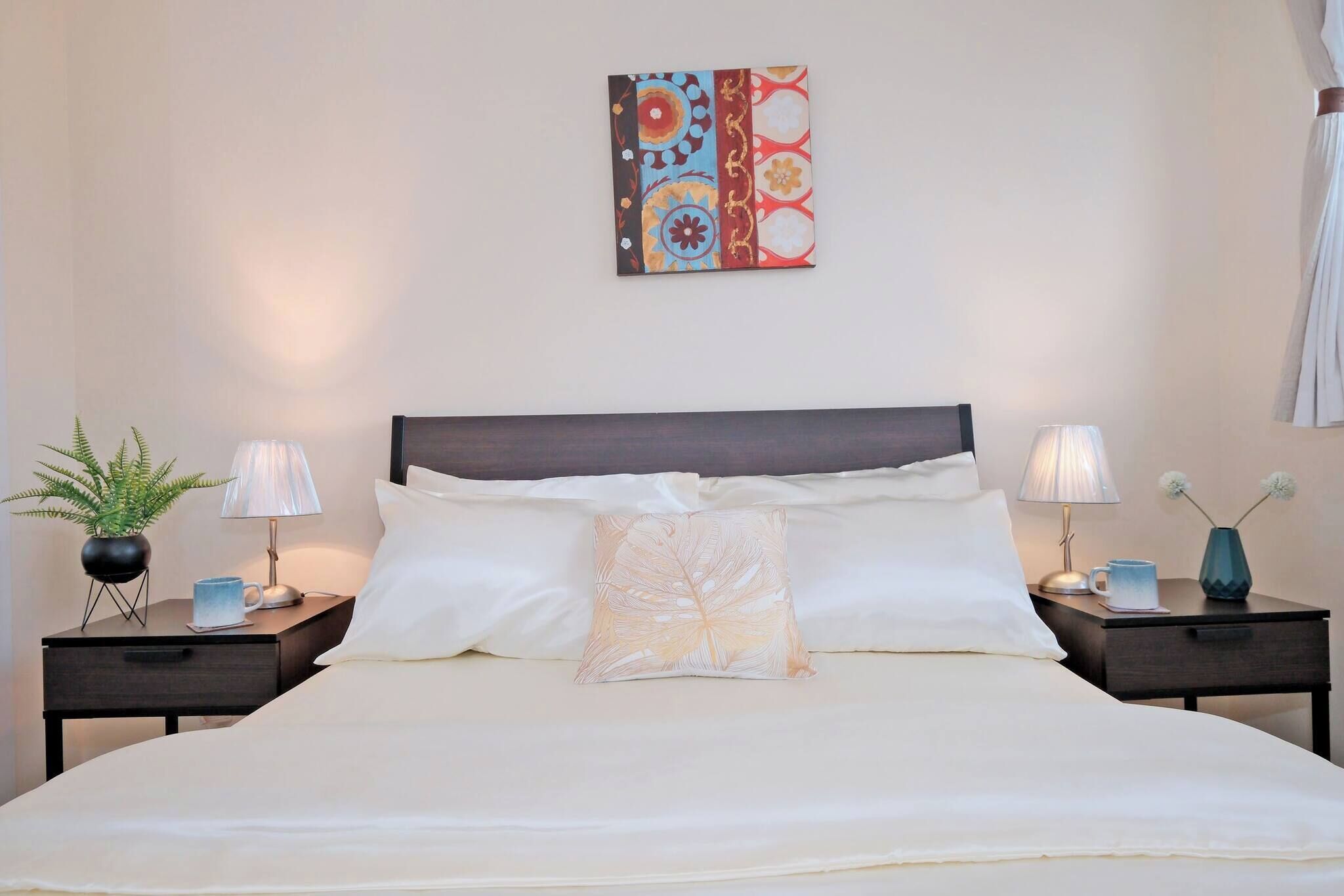 Een net opgemaakt bed met witte lakens, twee nachtkastjes met lampen, een decoratief kussen en een kunstwerk boven het bed.