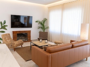 Una sala de estar moderna con un sofá de cuero, un sillón de mimbre, un televisor de pantalla plana montado en la pared, una chimenea y un ventanal con cortinas translúcidas.
