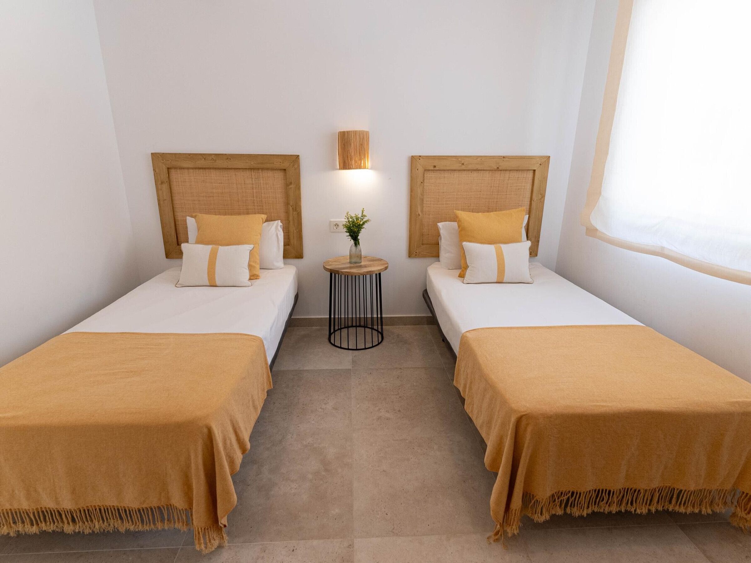 A hotel room with two beds, a small table with a lamp, and a window.