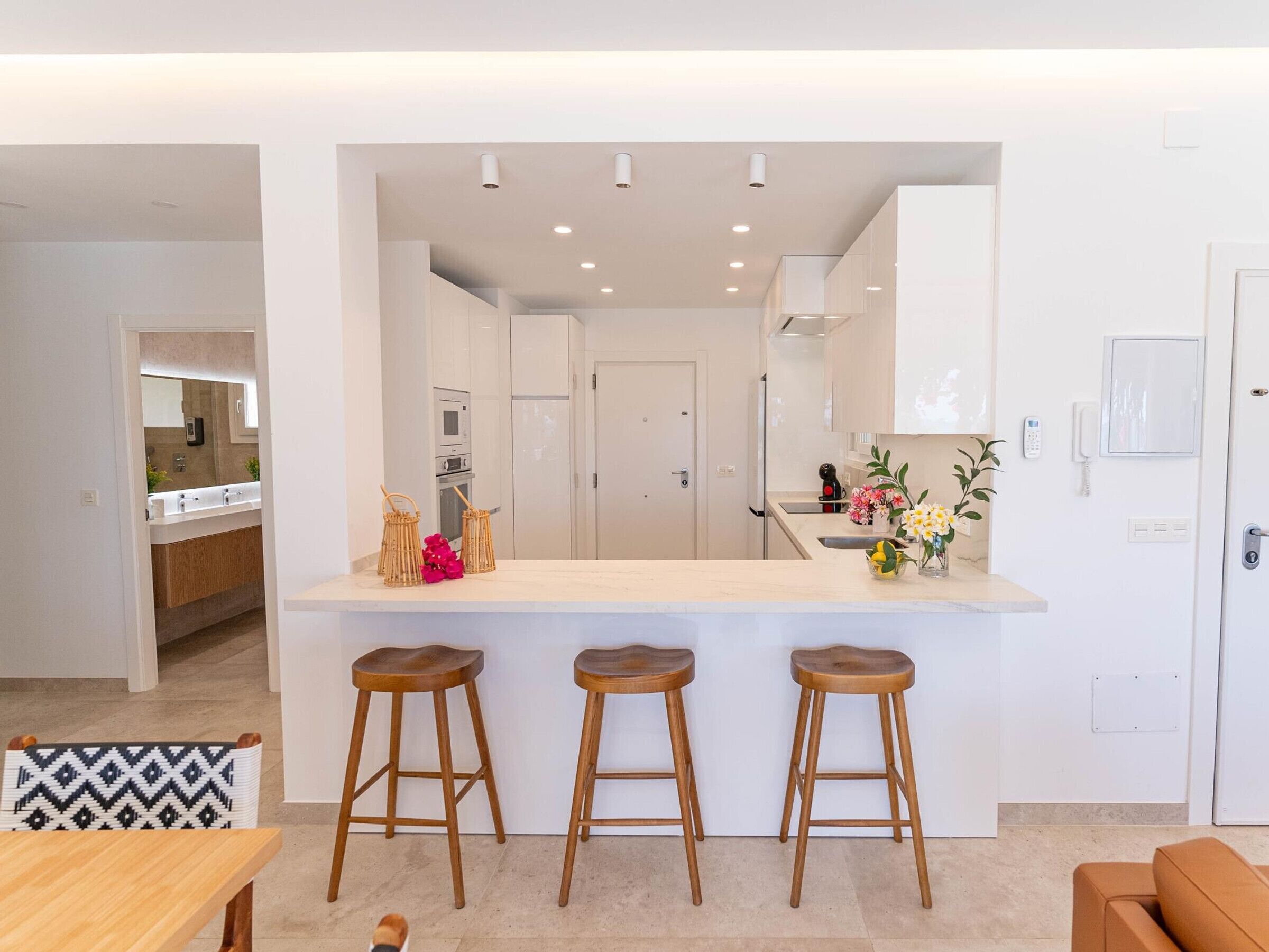 A modern kitchen with a central island, three wooden stools, and a dining area with a patterned chair.