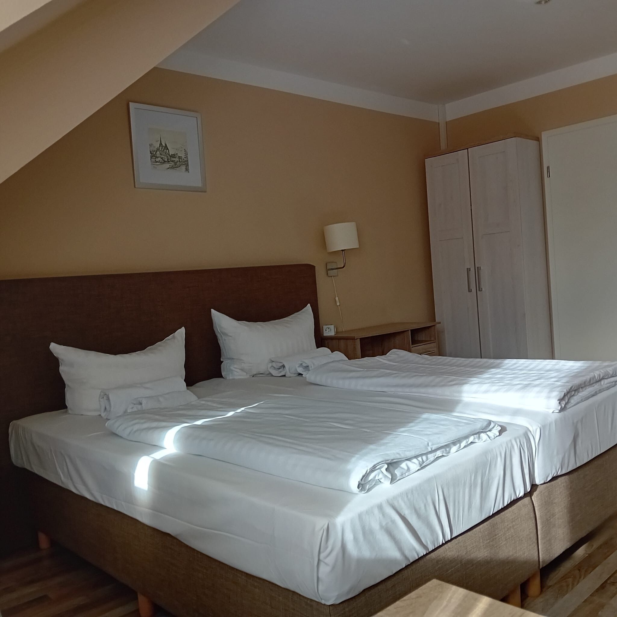 A neatly made bed in a room with a sloped ceiling, a wooden headboard, a wall-mounted lamp, and a wardrobe.
