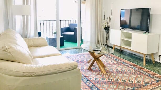 Ett vardagsrum med en soffa, ett soffbord i glas, en tv på ett tv-bord och en balkong med utemöbler.