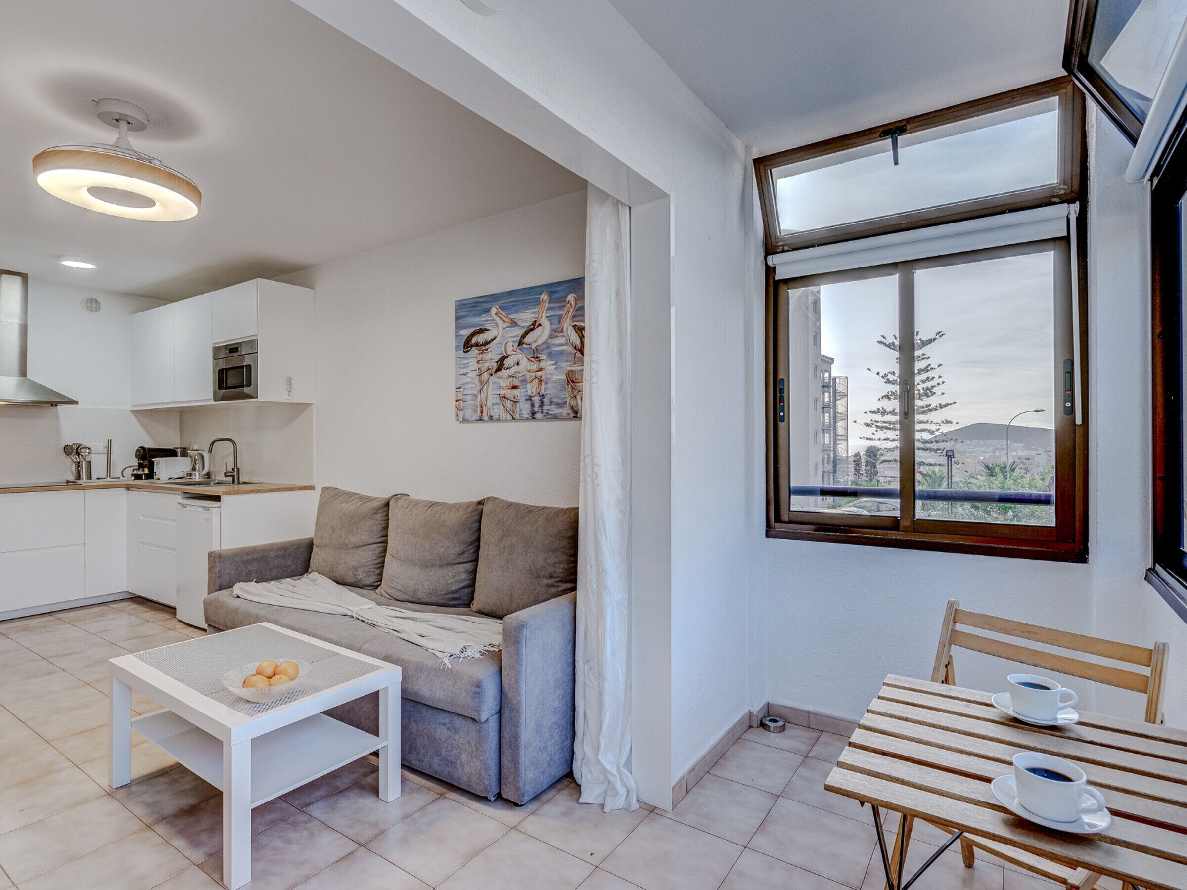 A compact living space with a kitchenette, a sofa, a dining table with cups, and a large window with a view.