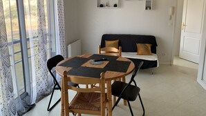 Ein Essbereich mit einem runden Holztisch, vier Stühlen und einer Couch mit Kissen. An der Wand befinden sich drei kleine Regale, eines mit der Aufschrift 'HOME'.