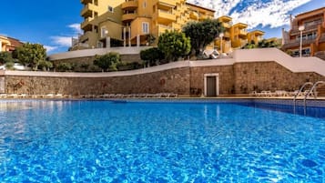 A swimming pool with clear blue water, surrounded by a stone wall and a yellow building with multiple balconies.