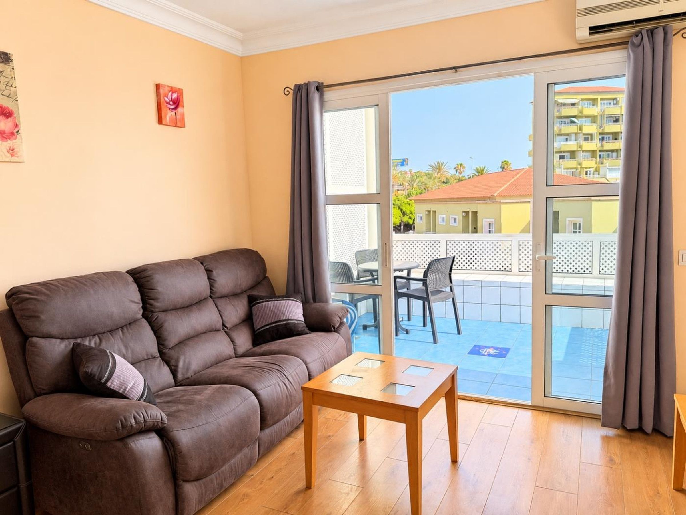 A living room with a brown sofa, a wooden coffee table, and a sliding glass door leading to an outdoor area with a balcony.