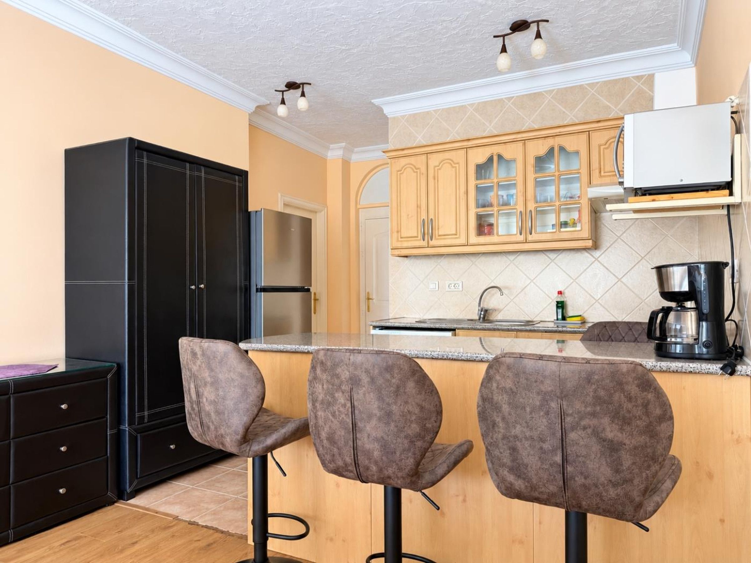 Una cocina con un refrigerador negro, armarios de madera, una cafetera y dos taburetes de bar.