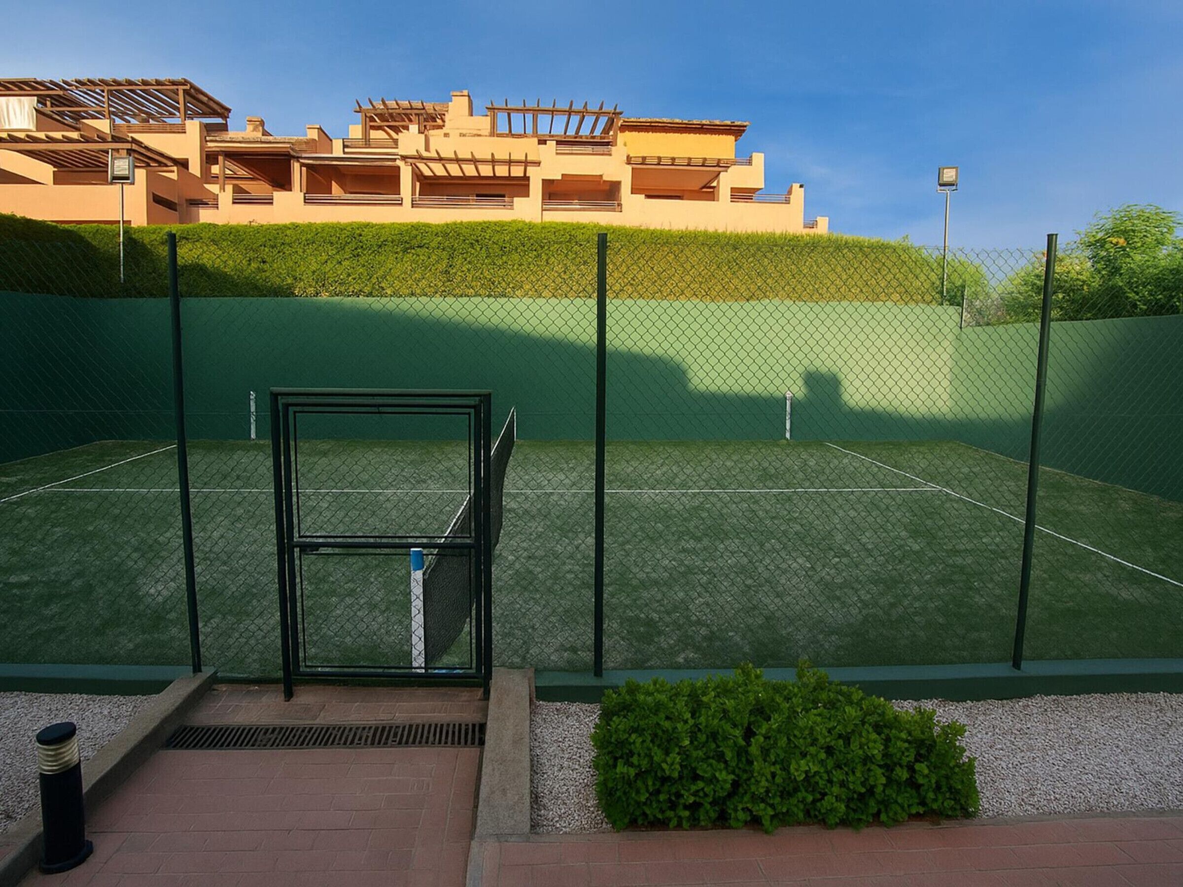 Una cancha de tenis con red, rodeada por una cerca verde y un seto.