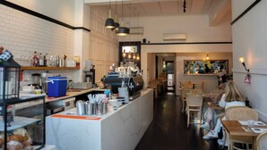 L’intérieur d’un café, avec un bar, des tables et un comptoir où sont exposés divers articles.