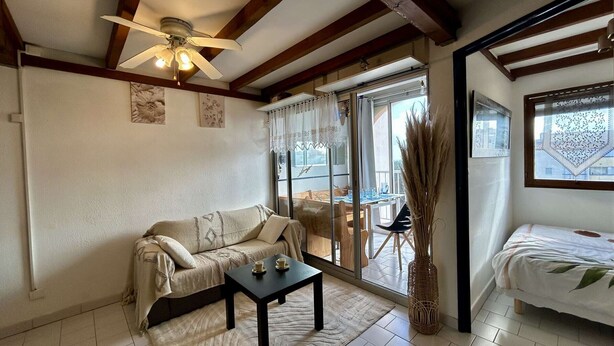 Una sala de estar acogedora con un sofá, una mesa de centro y un ventilador de techo. Se ve una ventana grande con vistas al exterior.