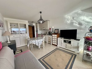 Ein Wohnzimmer mit Sofa, Esstisch, Fernseher und Küchenzeile mit Schränken und Kühlschrank.