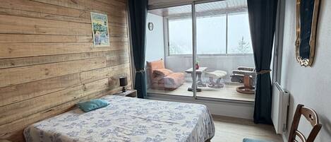 Ein Schlafzimmer mit einer Holzwand, einem Bett mit Blumenmuster-Bettwäsche, einem Sessel und Blick auf einen Balkon mit Gartenmöbeln.