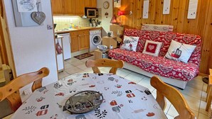 Ein gemütliches Wohnzimmer mit einer roten, gemusterten Couch, Holzwänden, einem Esstisch mit einer gemusterten Tischdecke und einer Küchenzeile mit Holzschränken und Geräten.