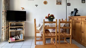 Un salon comprenant un téléviseur, des chaises en bois, une table agrémentée d’un centre de table floral et un meuble. Des drapeaux sont accrochés au mur.