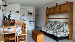 Un espace de vie compact comprenant un coin cuisine, un espace repas avec une table et des chaises, et un canapé à motifs tropicaux.