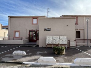 A building with a beige exterior, a red door, and a sign that reads "RESIDENCE LES JARDINS DE LA LAGUE." There are three white utility boxes in front of the building. - 2-Room Apartment with Parking in Banyuls-sur-Mer (Banyuls-sur-Mer)