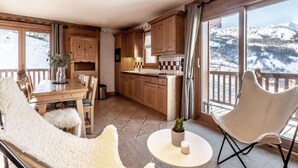 A cozy room with a wooden dining table, chairs, a small white table, and a view of snowy mountains through large windows. - Luxury Chalet: Sleeps 6, Fully Equipped, Near Slopes (Saint-Martin-de-Belleville)