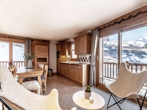 Een knus ingerichte kamer met een houten eettafel, stoelen, een klein witte tafeltje en uitzicht op besneeuwde bergen door grote ramen.