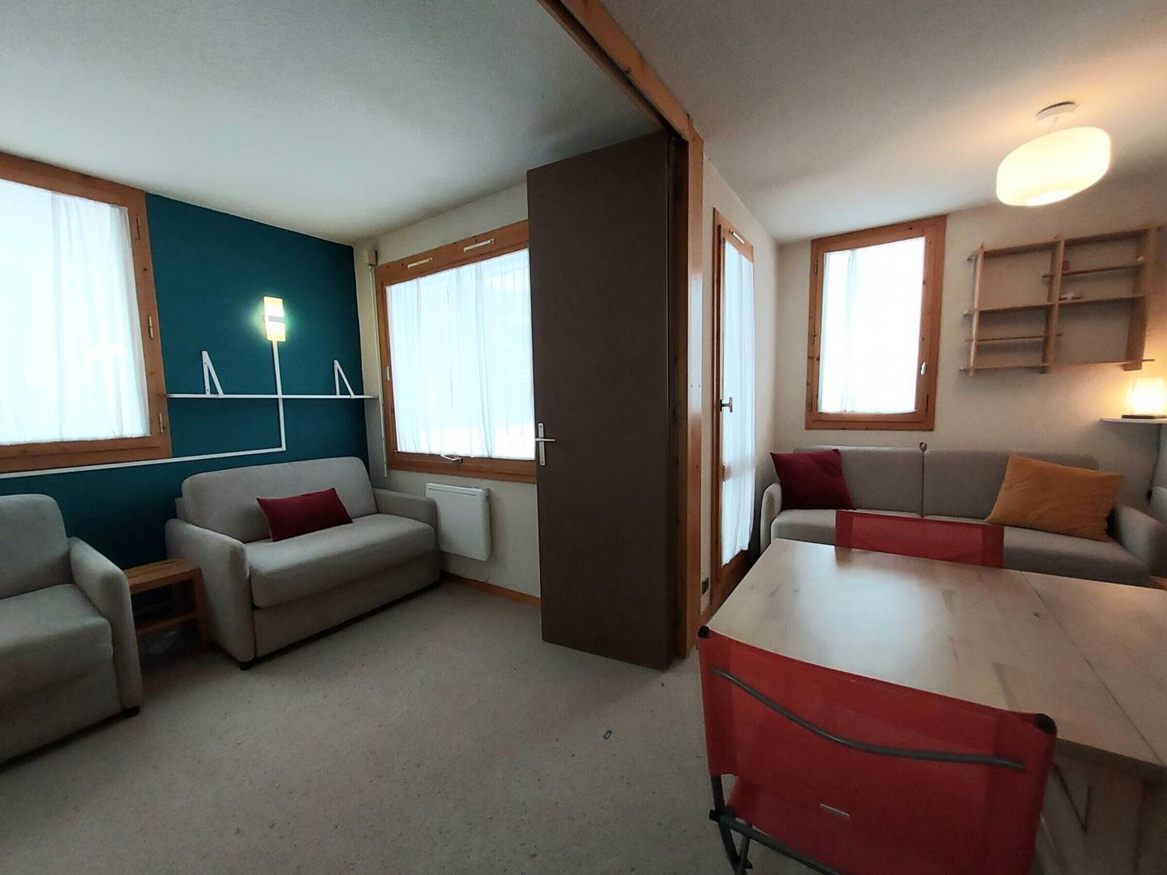 A room with two sofas, a table, and a red chair. There are windows with curtains, a lamp, and a shelf with items.