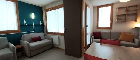 A room with two sofas, a table, and a red chair. There are windows with curtains, a lamp, and a shelf with items.
