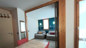 A room with a wooden dining table, two sofas, and a red chair. There is a wooden door frame and a window with curtains. - Cozy Studio on the Slopes - Sleeps 4 (Valmorel)