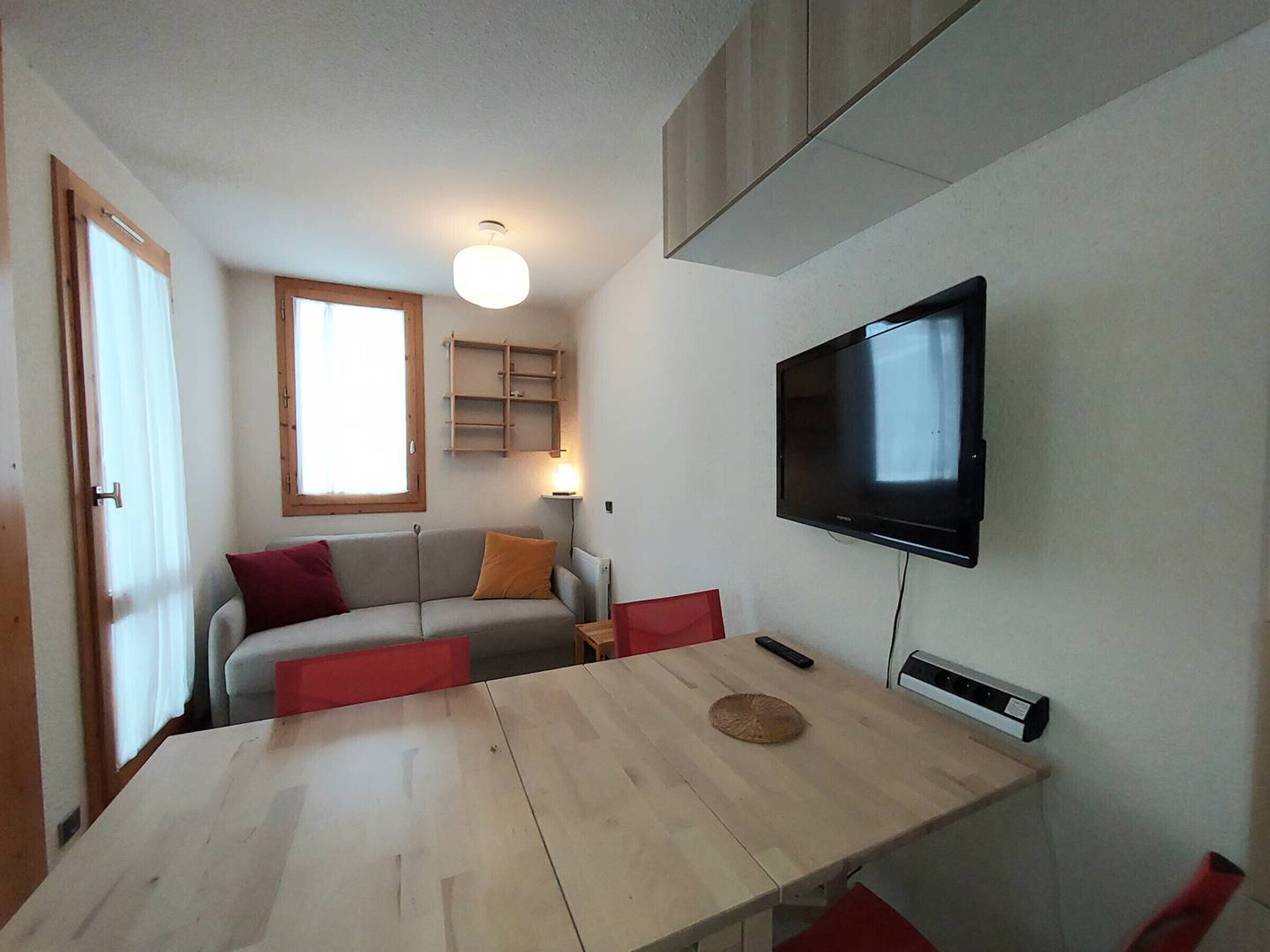 A compact living space with a sofa, a dining table, a TV mounted on the wall, and a window providing natural light.