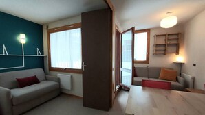 A modern living room with a sofa, a dining table, and a large window. - Cozy Studio on the Slopes - Sleeps 4 (Valmorel)