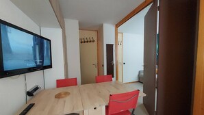 A room with a TV mounted on the wall, a wooden table with red chairs, and a door leading to another room. - Cozy Studio on the Slopes - Sleeps 4 (Valmorel)