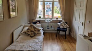 A small, cozy room with two beds, a small table, and a chair. There is a window with curtains, a radiator, and a door. - Room-walkinstown Retreat House (Dublin)