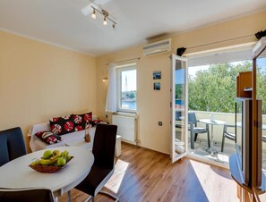 Ein modernes Wohnzimmer mit Essbereich, einem Sofa mit gemusterten Kissen, einem Tisch mit Früchten und einem Balkon mit Gartenmöbeln.