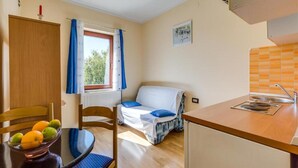 Ein kleines Zimmer mit Bett, einem Tisch mit Obst, einem mit Vorhängen versehenen Fenster, einem Heizkörper und einer Küchenzeile.