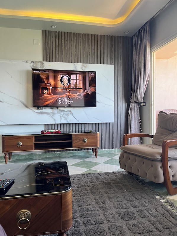 A living room with a flat-screen TV mounted on a marble wall, a wooden entertainment unit, a tufted leather armchair, and a patterned rug. - seaview hotel (Alexandria)