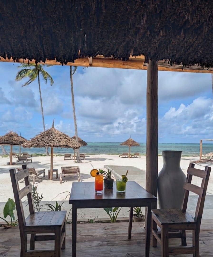 En restaurang vid stranden med parasoller av halm, träbord och stolar, med utsikt över havet.