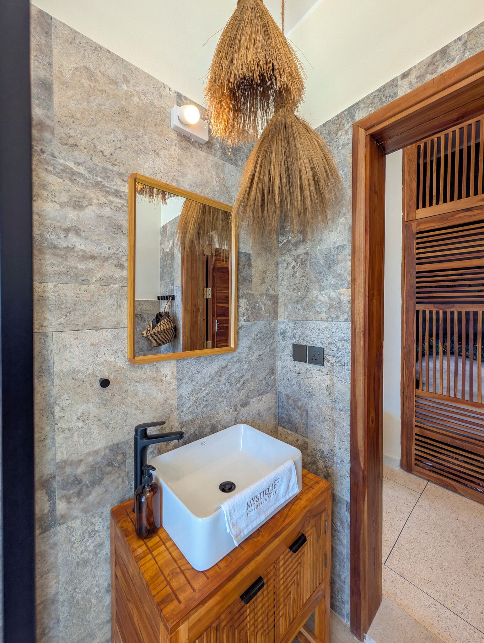 A bathroom with a stone wall, a white sink, a wooden vanity, a mirror, and a hanging light fixture.