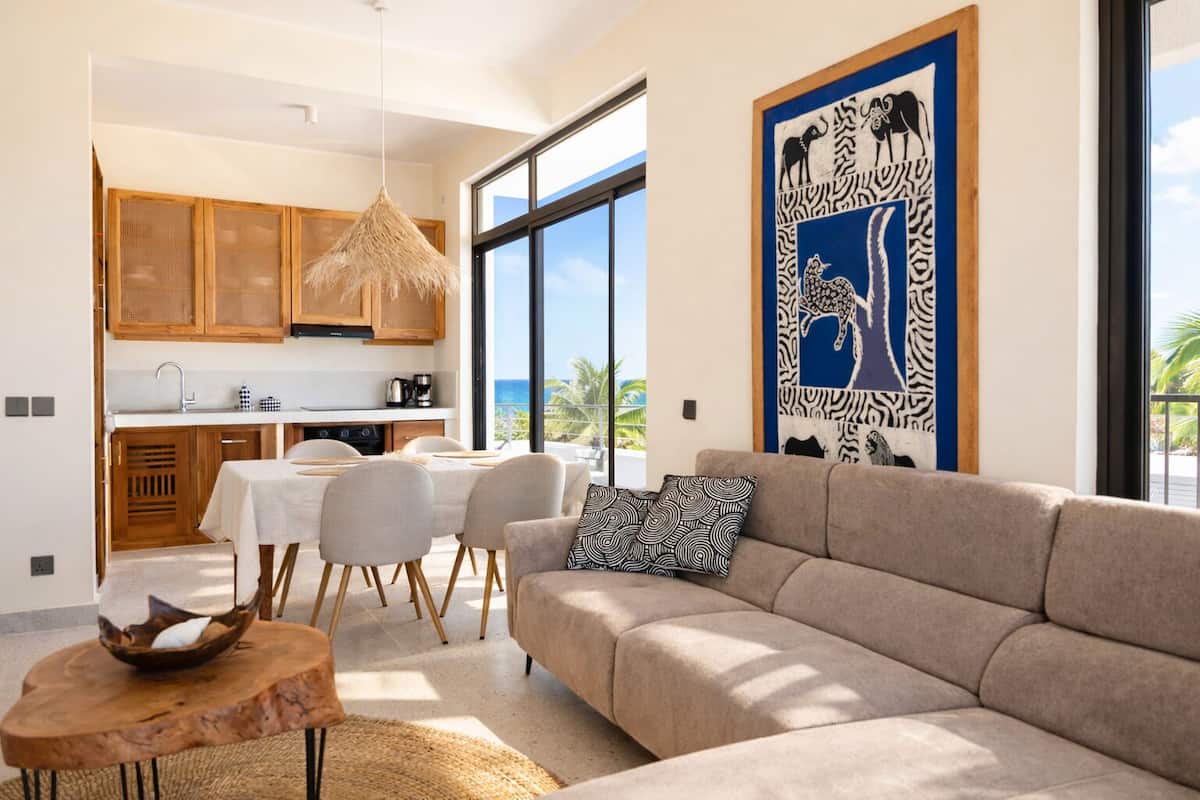 Eine moderne Küche mit Holzausstattung, ein Essbereich mit Tisch und Stühlen, ein Sofa mit einem gemusterten Kissen und ein großes Fenster mit Meerblick.