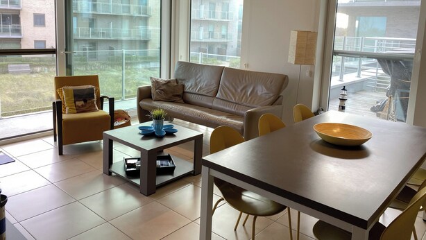 Una sala de estar moderna con sofá, sillones, una mesa de centro y una mesa de comedor. Amplias ventanas ofrecen vistas al exterior.