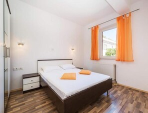 A modern bedroom with a wooden floor, a bed with white linens and orange pillows, a nightstand, and a window with orange curtains. - 1-bedroom Sea-view Apartment in Obrovac (Obrovac)