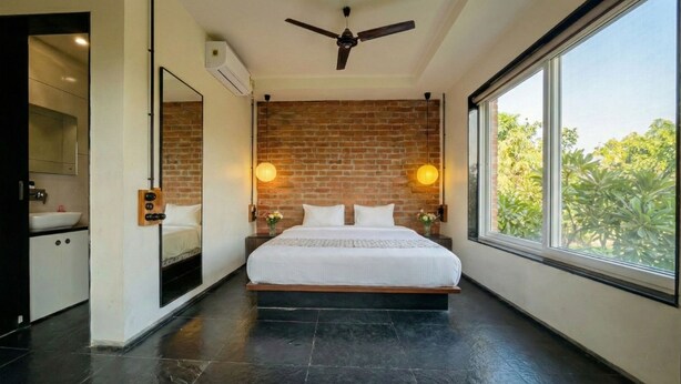 A modern bedroom with a brick wall, a large bed, a ceiling fan, and a window with a view of greenery. - Brick Villa Heritage  (Trimbak)