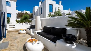 Uma área de estar externa com piso de pedra, um sofá branco com almofadas pretas e uma mesa com comida e bebidas.