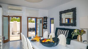 Uma sala de estar moderna com sofá, mesa de jantar com frutas e bebidas, espelho e vista através das portas de vidro.