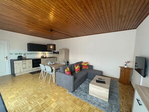 Een moderne keuken met een eetgedeelte, een grijze bank met kleurrijke kussens, en een houten plafond.