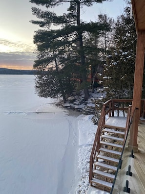 A wooden deck with a staircase leading to a covered area, surrounded by snow-covered trees and a frozen lake.