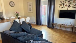 Uma sala de estar moderna com um sofá escuro, uma mesa de jantar com cadeiras, uma televisão de tela plana na parede e uma janela com cortinas.