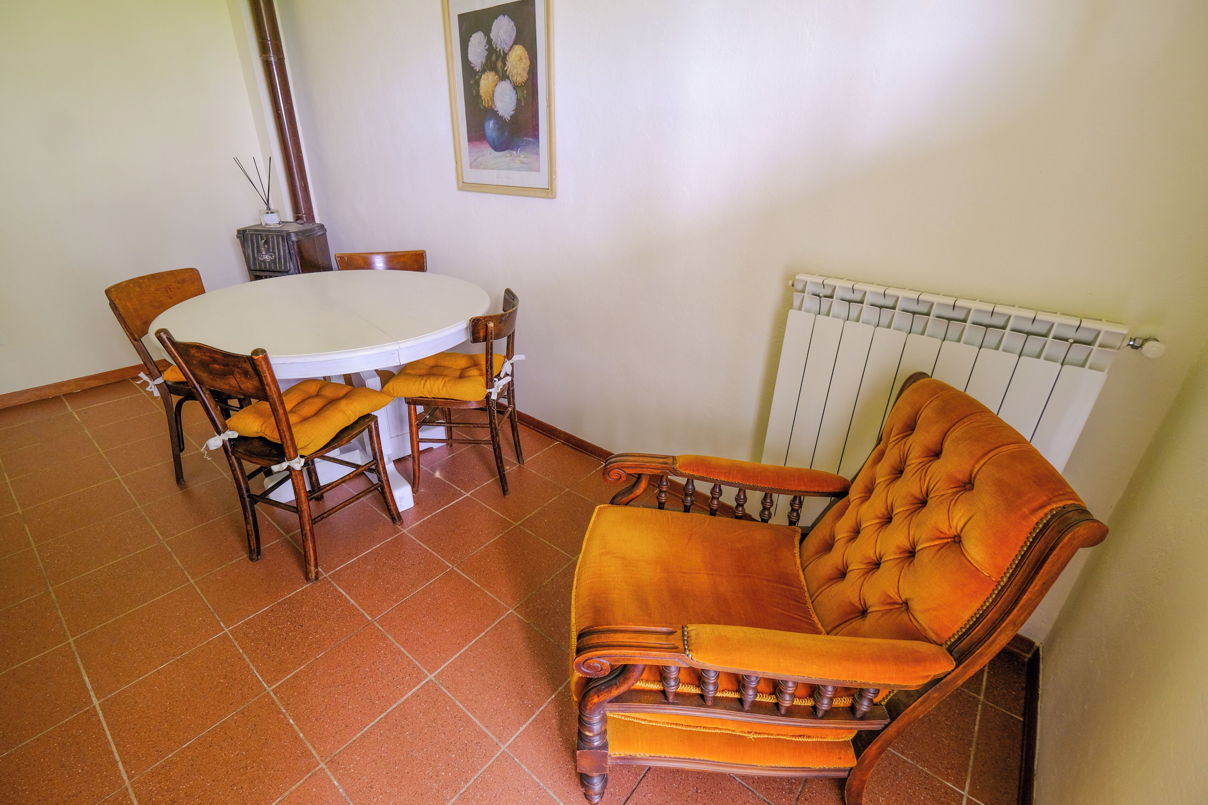 Una zona de menjador amb una taula rodona i quatre cadires, un sofà, un radiador i un quadre amb marc a la paret.